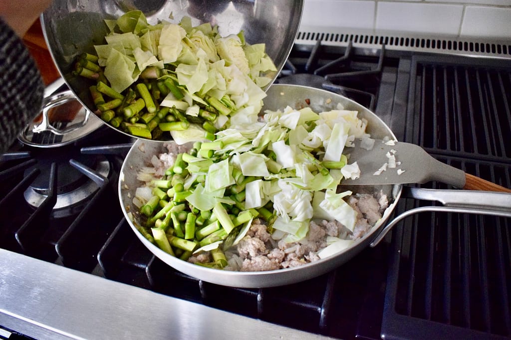 Adding chopped vegetables from a large bowl to skillet of pork stir fry.