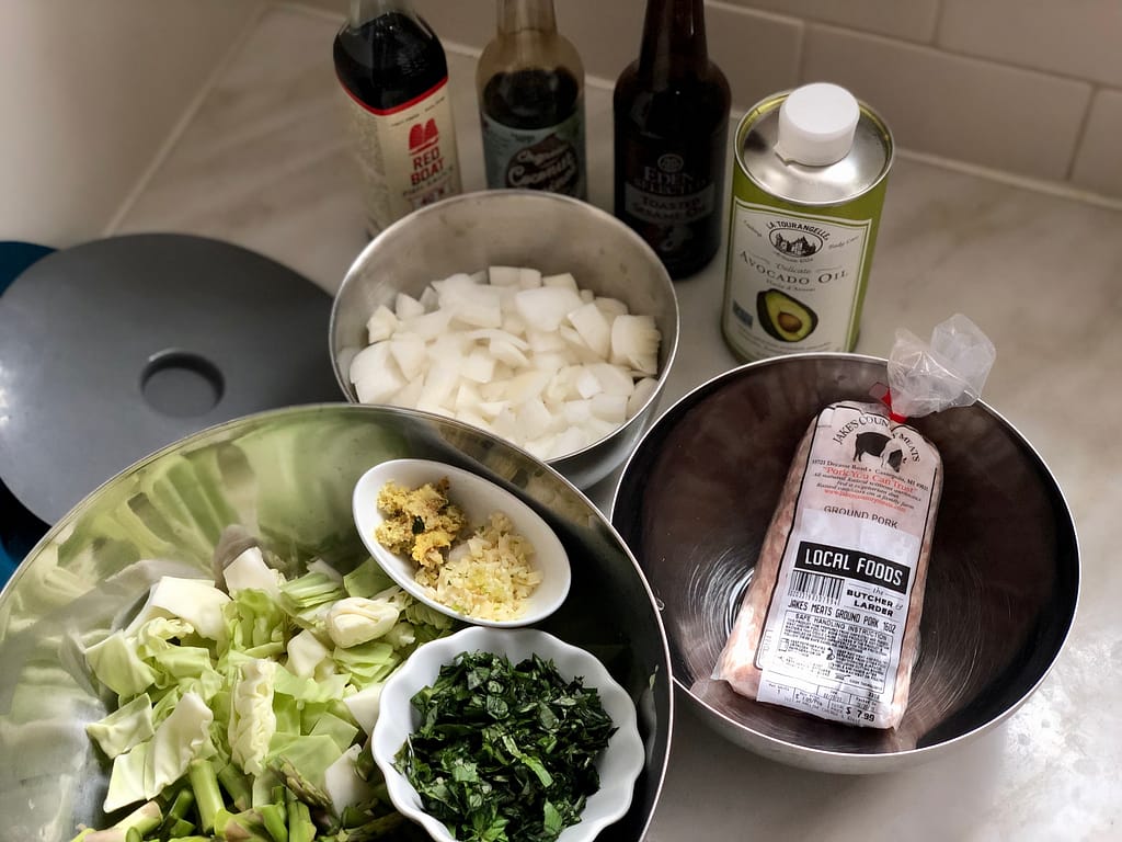 All ingredients prepped and arranged on counter top for veggie loaded pork stir fry. Bowl of cabbage, asparagus, ginger, garlic, basil, chopped onion, ground pork and sauces.