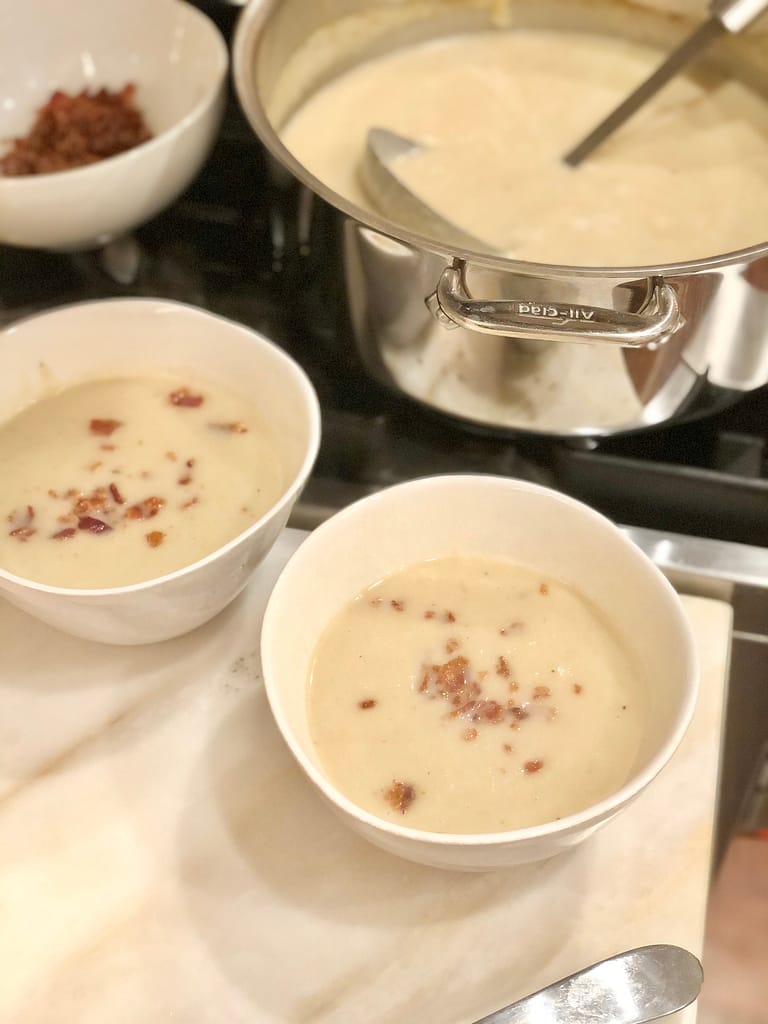 Pot of cauliflower soup with two bowls of soup, side of bacon and a woman holding Hepp's truffle salt for garnish.Pot of cauliflower soup with two bowls of soup with crumbled bacon.