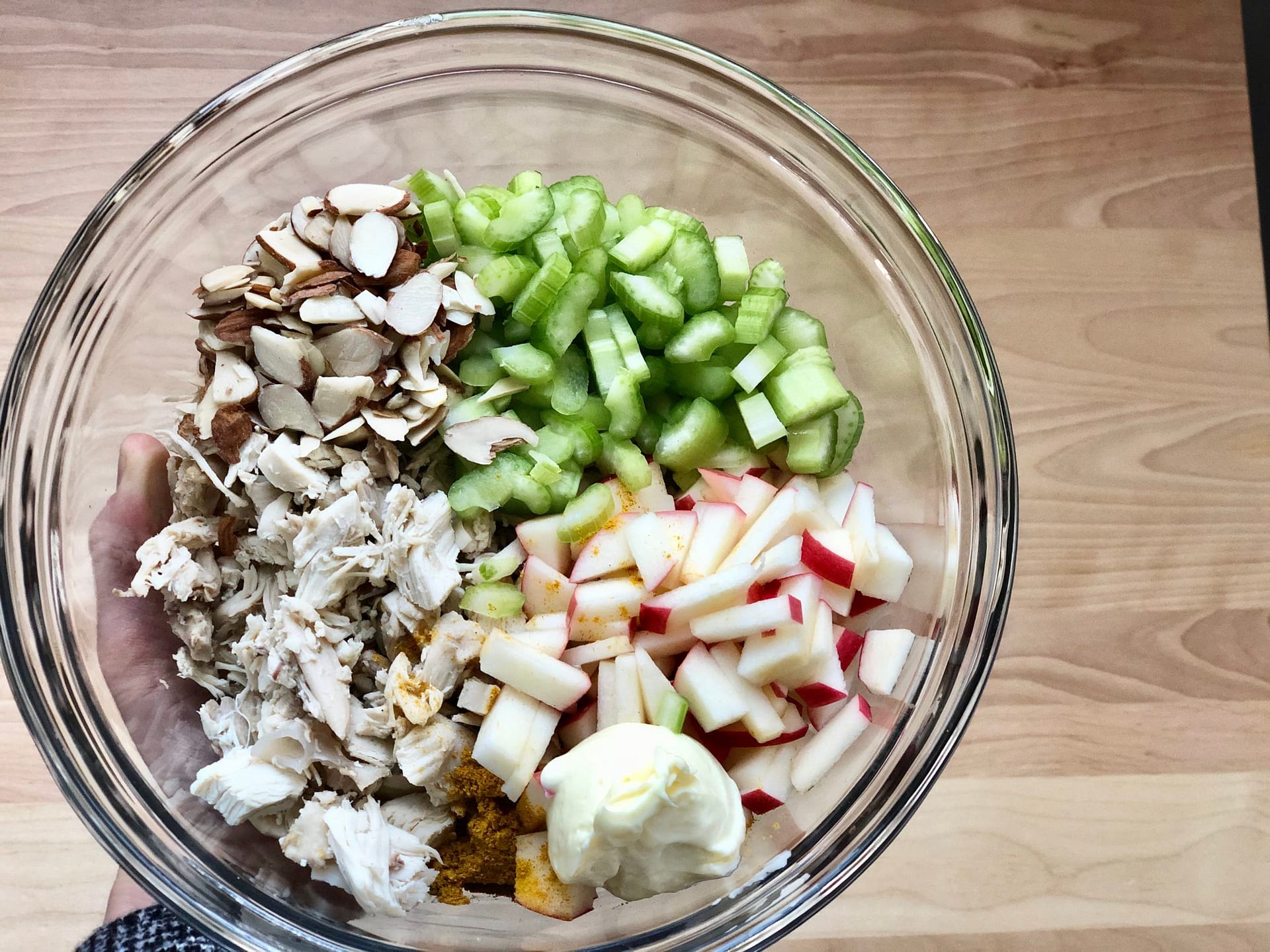 Glass bowl with ingredients for healthy curry chicken salad. Chopped apple, chopped celery, sliced almonds, cooked and chopped chicken, curry powder, and avocado oil mayonnaise.